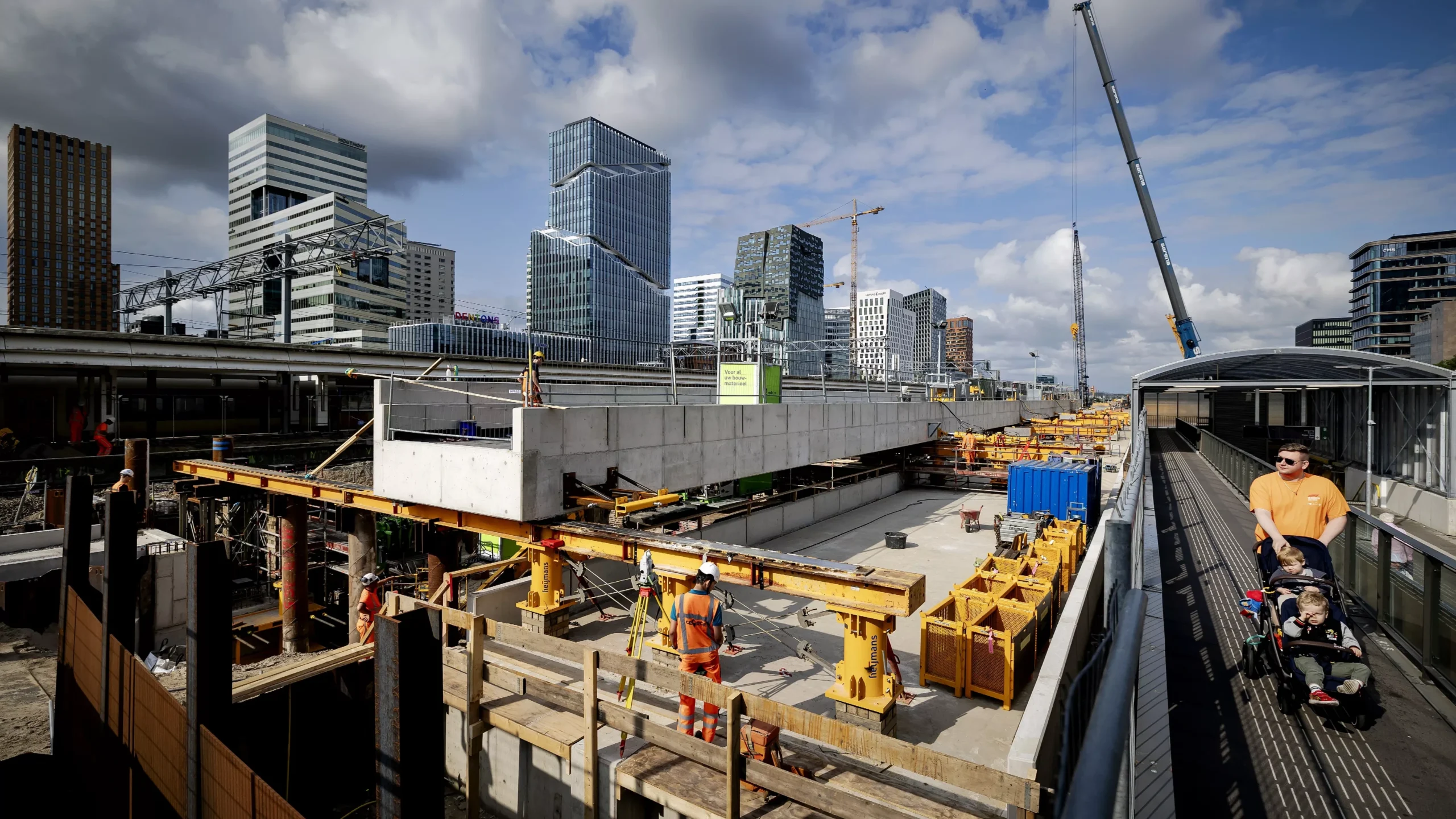Bouwplaats in een moderne skyline met kranen, werknemers en voorbijlopende mensen op een loopbrug.