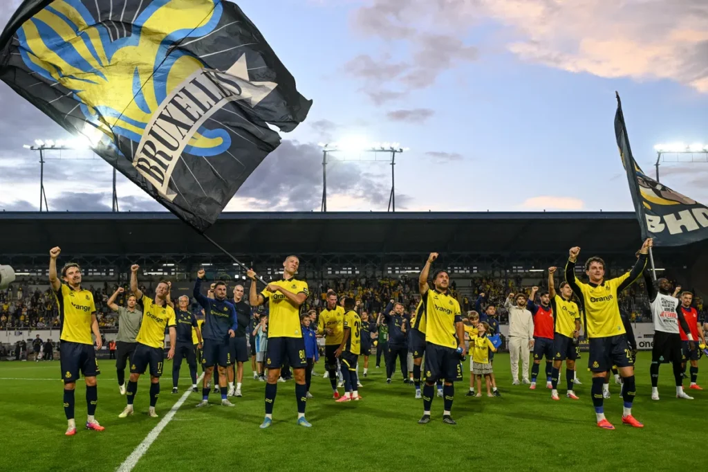 Union Saint-Gilloise spelers en supporters op het veld na een wedstrijd, met grote clubvlaggen in het stadion.