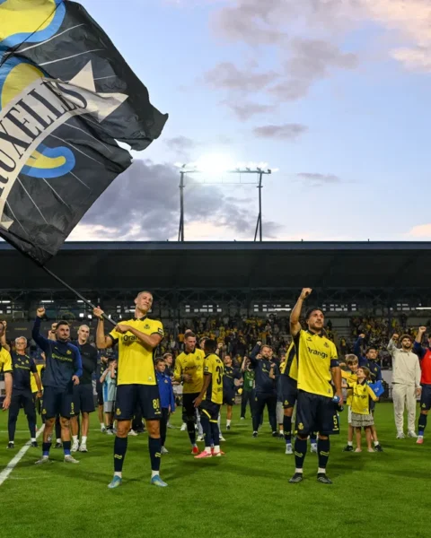 Union Saint-Gilloise spelers en supporters op het veld na een wedstrijd, met grote clubvlaggen in het stadion.