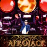 Afrojack staat op het podium tijdens een optreden, met band en camera’s om hem heen.