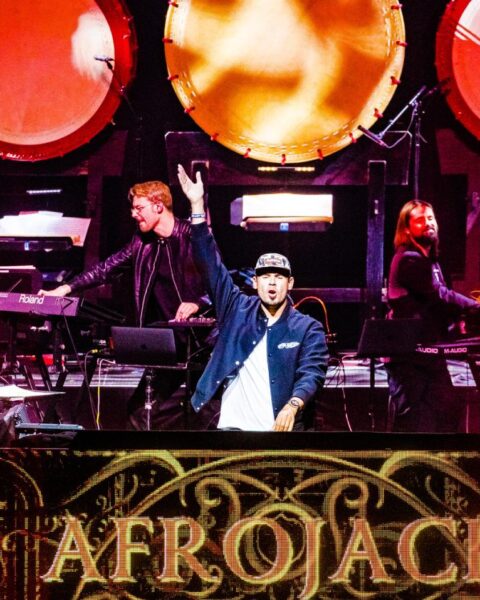 Afrojack staat op het podium tijdens een optreden, met band en camera’s om hem heen.