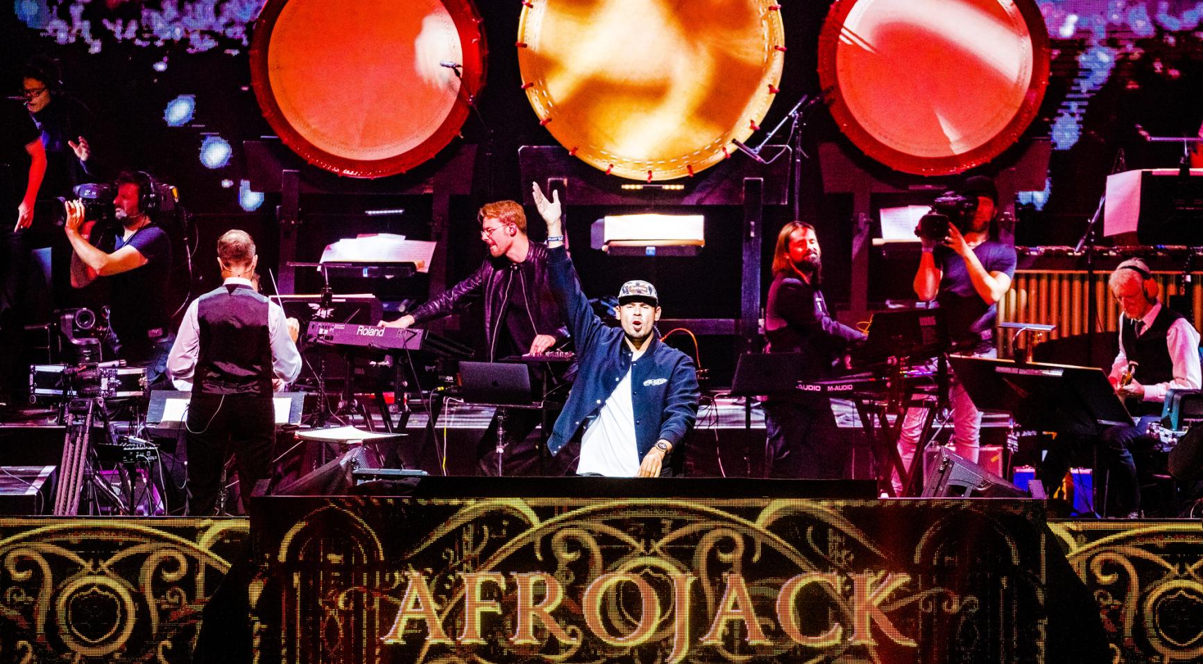 Afrojack staat op het podium tijdens een optreden, met band en camera’s om hem heen.