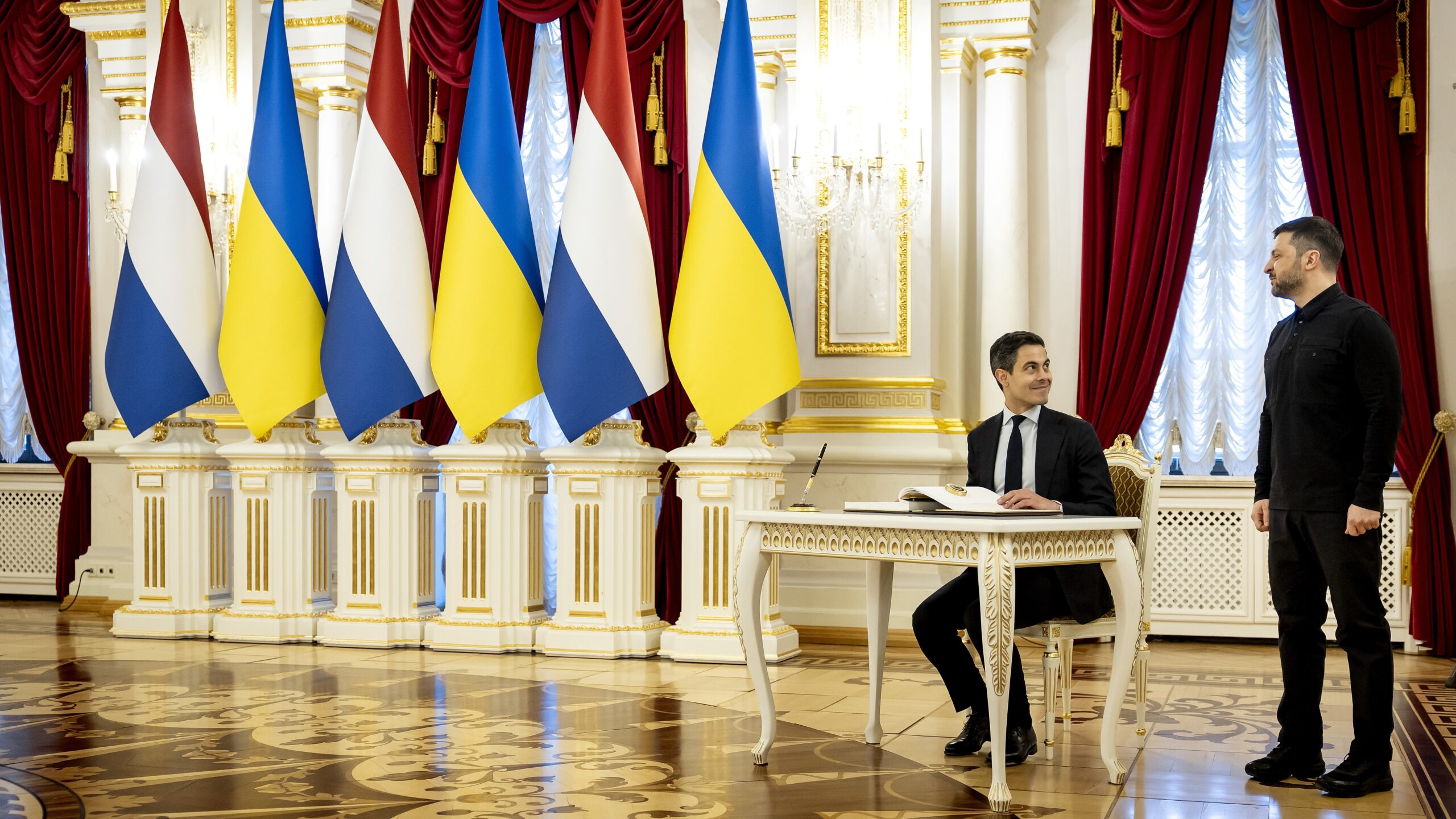Volodymyr Zelensky zit aan een tafel in een rijkversierde zaal met Oekraïense en Nederlandse vlaggen.