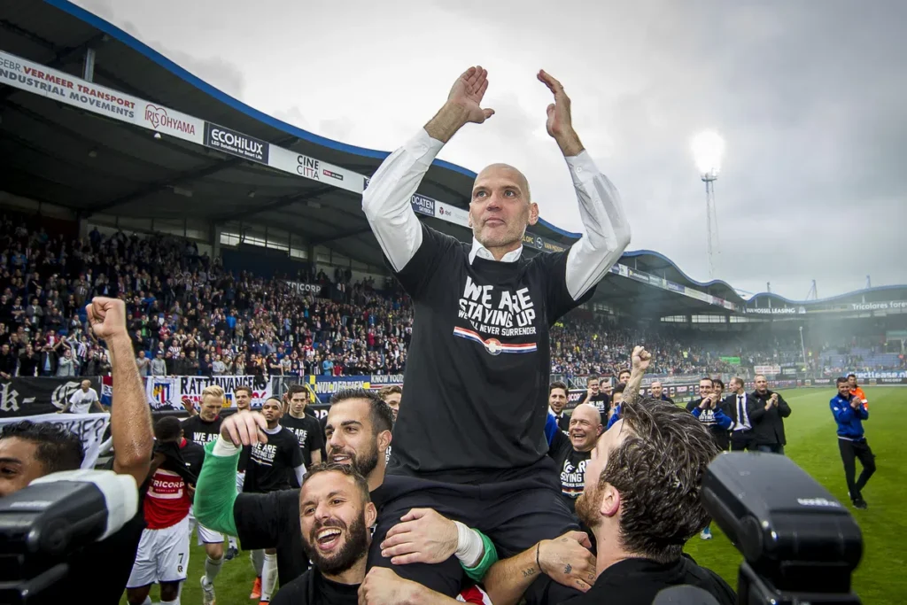 Jurgen Streppel wordt op het veld in een stadion gejuicht terwijl hij zijn armen omhoog steekt.
