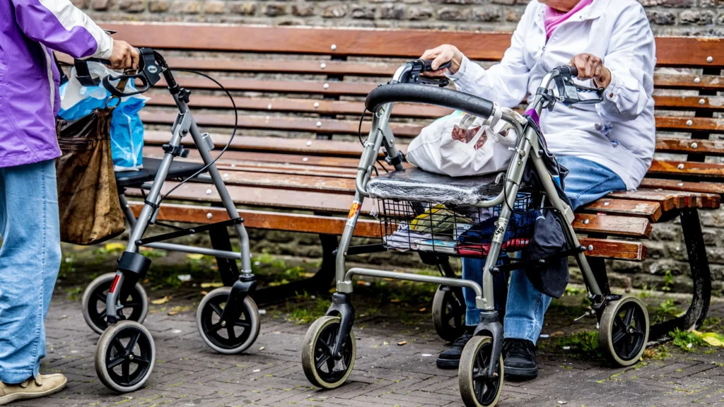 Twee oudere vrouwen met rollators op een bankje in een straatomgeving.