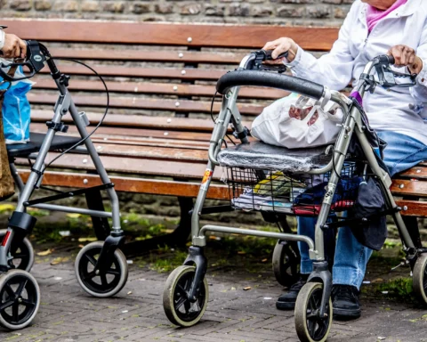 Twee oudere vrouwen met rollators op een bankje in een straatomgeving.