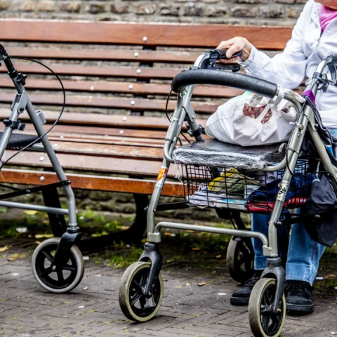 Twee oudere vrouwen met rollators op een bankje in een straatomgeving.