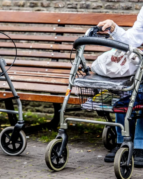 Twee oudere vrouwen met rollators op een bankje in een straatomgeving.