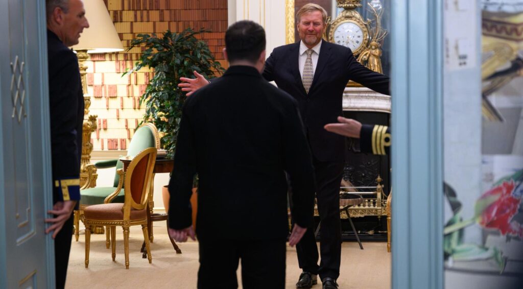 King Willem-Alexander staat met open armen in een ornamentele zaal terwijl een bezoeker hem nadert.