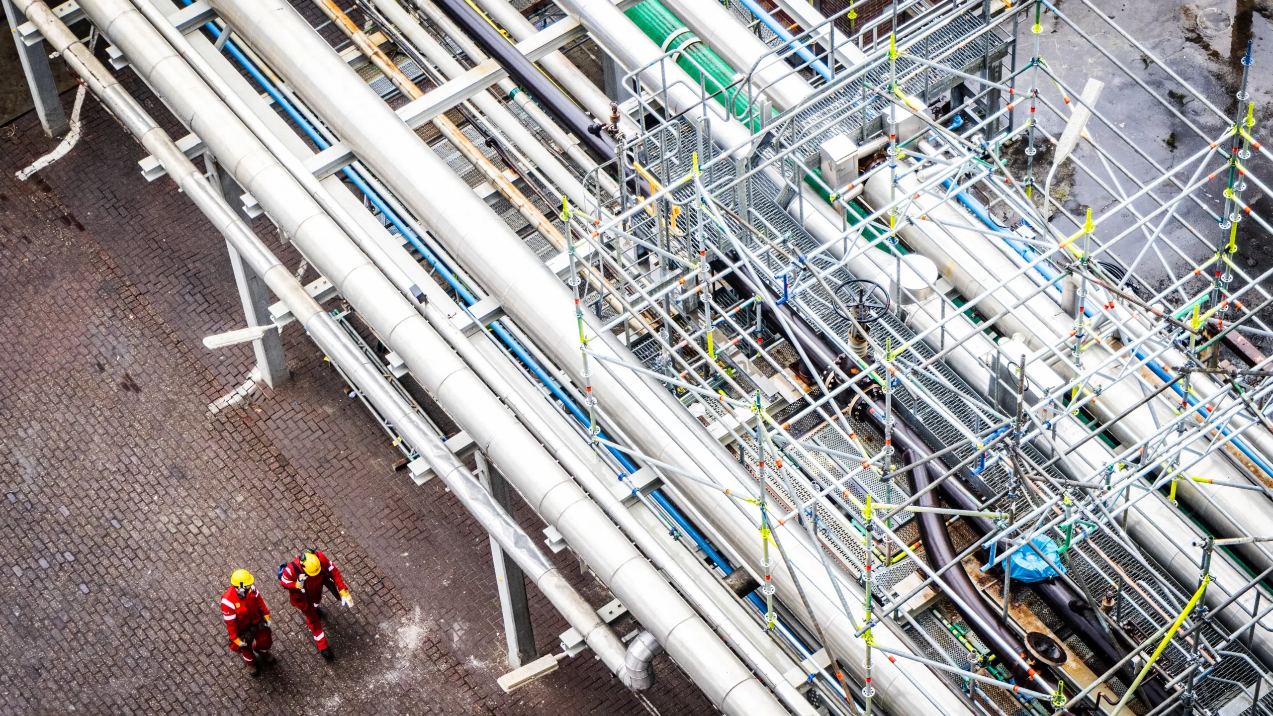 Luchtfoto van grote industriële pijpleidingen met twee werknemers in beschermende kleding eronder.
