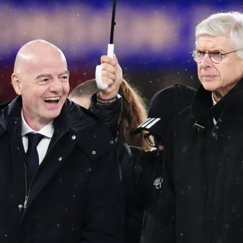 Gianni Infantino en Arsène Wenger staan naast elkaar in een stadion tijdens regenachtig weer.