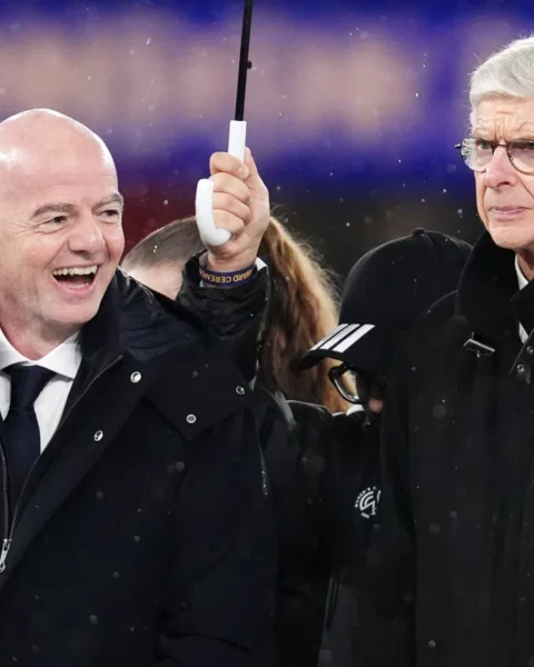 Gianni Infantino en Arsène Wenger staan naast elkaar in een stadion tijdens regenachtig weer.
