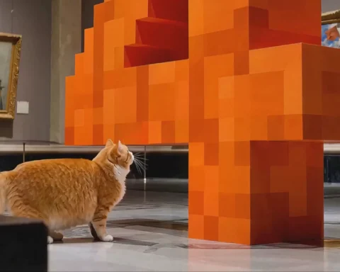 Een dikke oranje-witte kat staat naast een grote oranje pixelachtige sculptuur in een museumzaal.