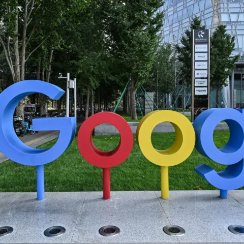Groot Google-logo bij een kantoorcampus met bomen en een moderne glazen gevel op de achtergrond.