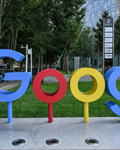 Groot Google-logo bij een kantoorcampus met bomen en een moderne glazen gevel op de achtergrond.