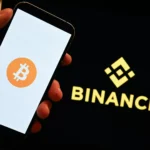 Hand houdt een telefoon vast met het Bitcoin-logo, naast een scherm met het Binance-logo.