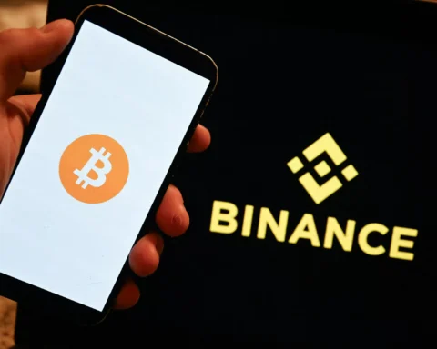 Hand houdt een telefoon vast met het Bitcoin-logo, naast een scherm met het Binance-logo.