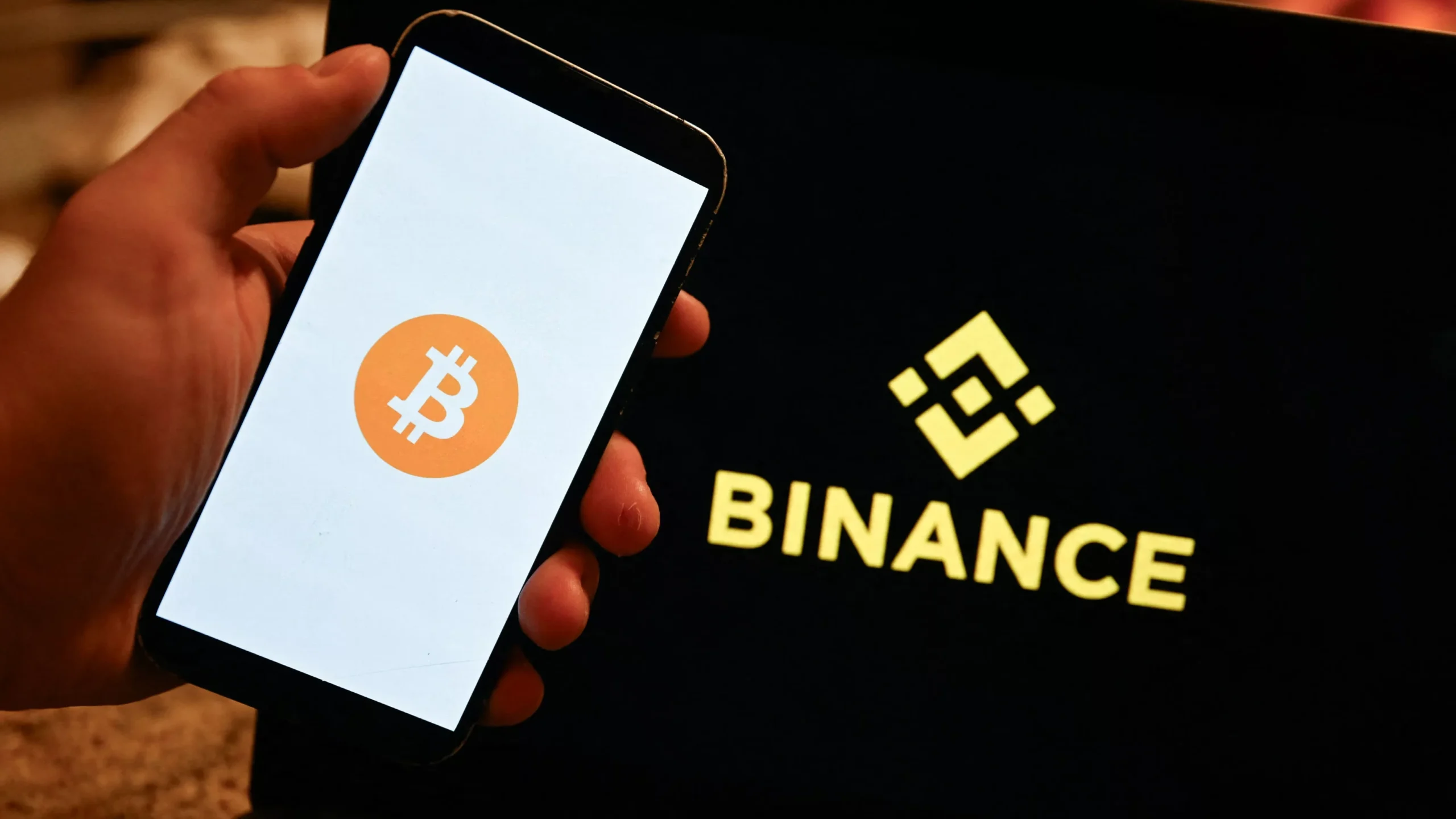 Hand houdt een telefoon vast met het Bitcoin-logo, naast een scherm met het Binance-logo.