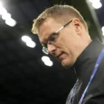 Een man met bril en zwarte jas staat in een stadion en kijkt omlaag.