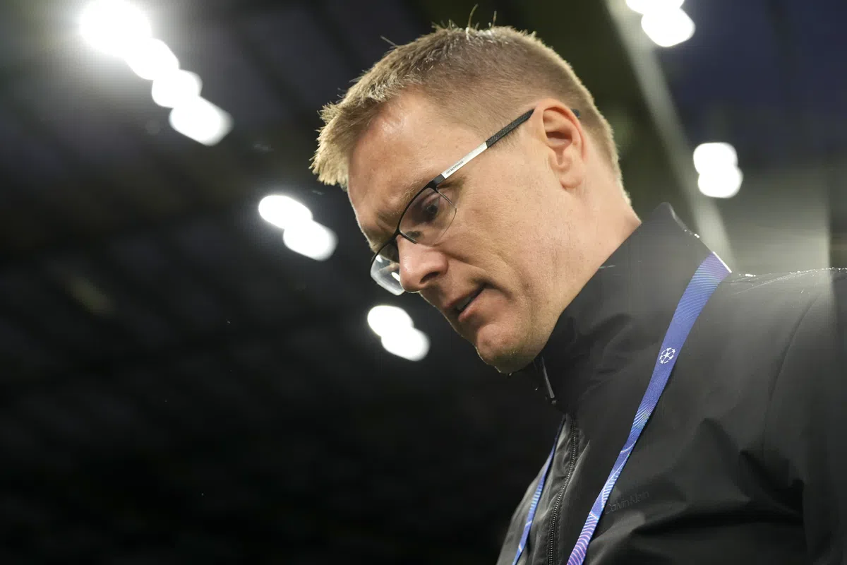 Een man met bril en zwarte jas staat in een stadion en kijkt omlaag.