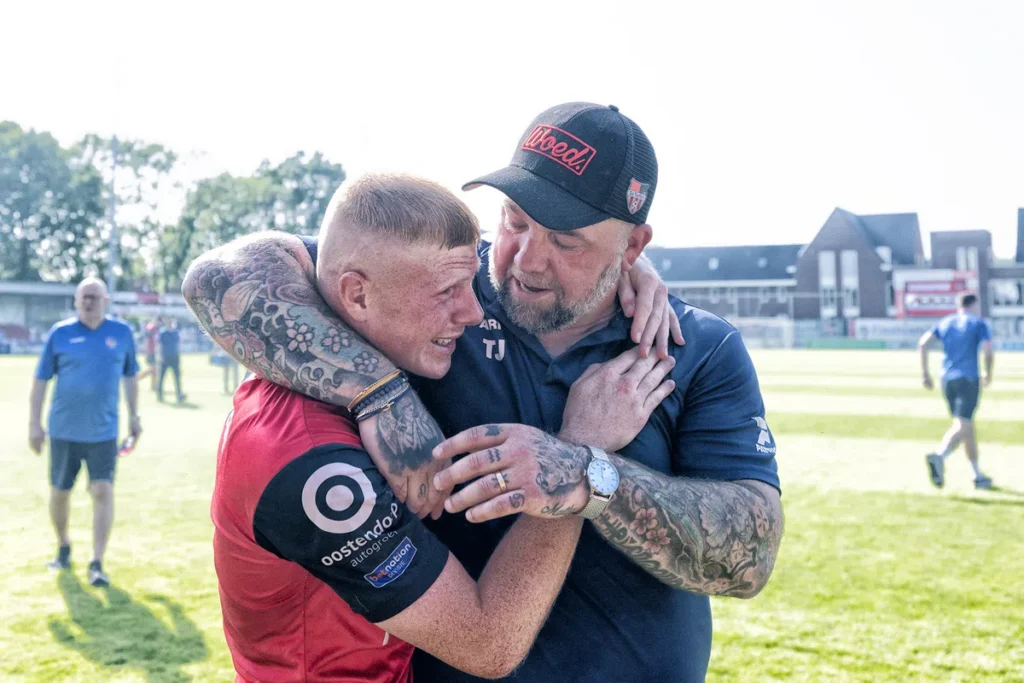 Een voetballer in rood shirt omhelst een trainer op het veld na een wedstrijd.