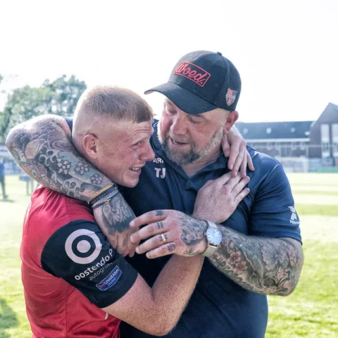 Een voetballer in rood shirt omhelst een trainer op het veld na een wedstrijd.