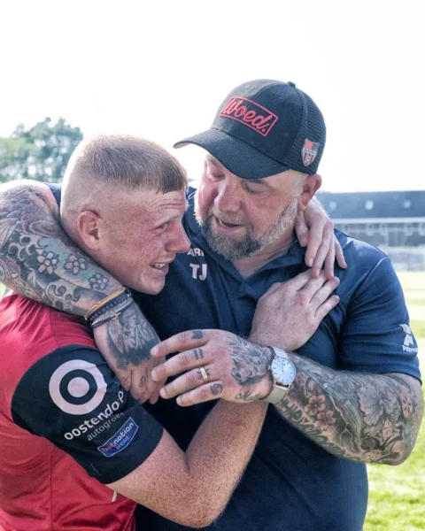 Een voetballer in rood shirt omhelst een trainer op het veld na een wedstrijd.