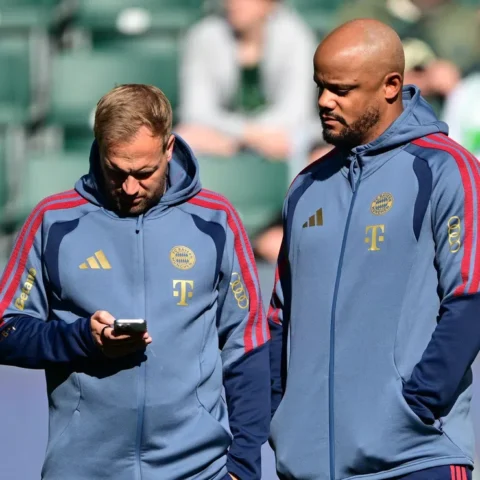 Twee Bayern München-trainers in clubtrainingspakken op het trainingsveld, één kijkt op een telefoon.