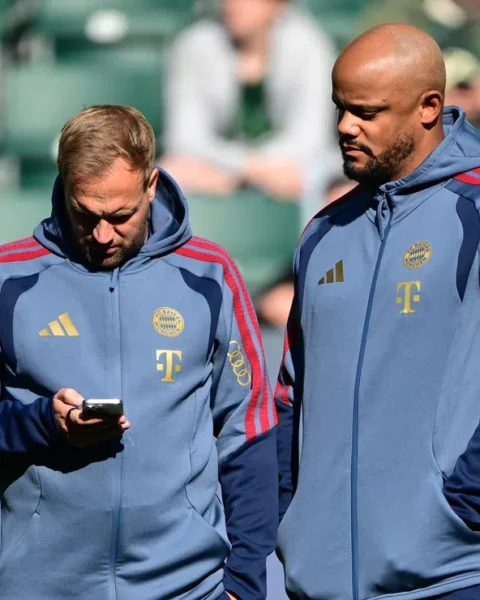 Twee Bayern München-trainers in clubtrainingspakken op het trainingsveld, één kijkt op een telefoon.