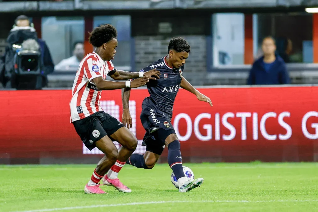 Twee voetballers in duel tijdens een wedstrijd, met een speler in rood-witte strepen die druk zet op een speler in donker tenue.