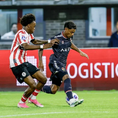 Twee voetballers in duel tijdens een wedstrijd, met een speler in rood-witte strepen die druk zet op een speler in donker tenue.