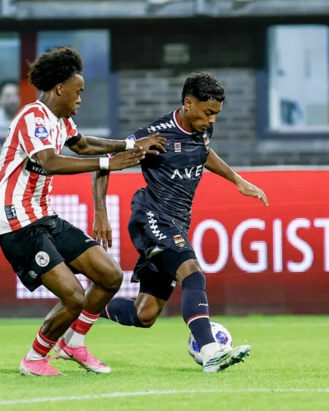 Twee voetballers in duel tijdens een wedstrijd, met een speler in rood-witte strepen die druk zet op een speler in donker tenue.
