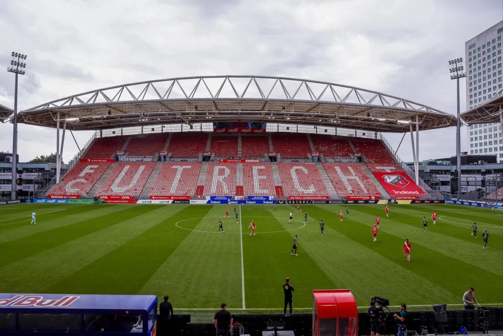 Een voetbalwedstrijd in Stadion Galgenwaard met lege rood-witte tribunes en spelers op het veld.