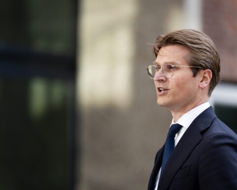 Een man in pak en bril staat buiten en kijkt opzij.