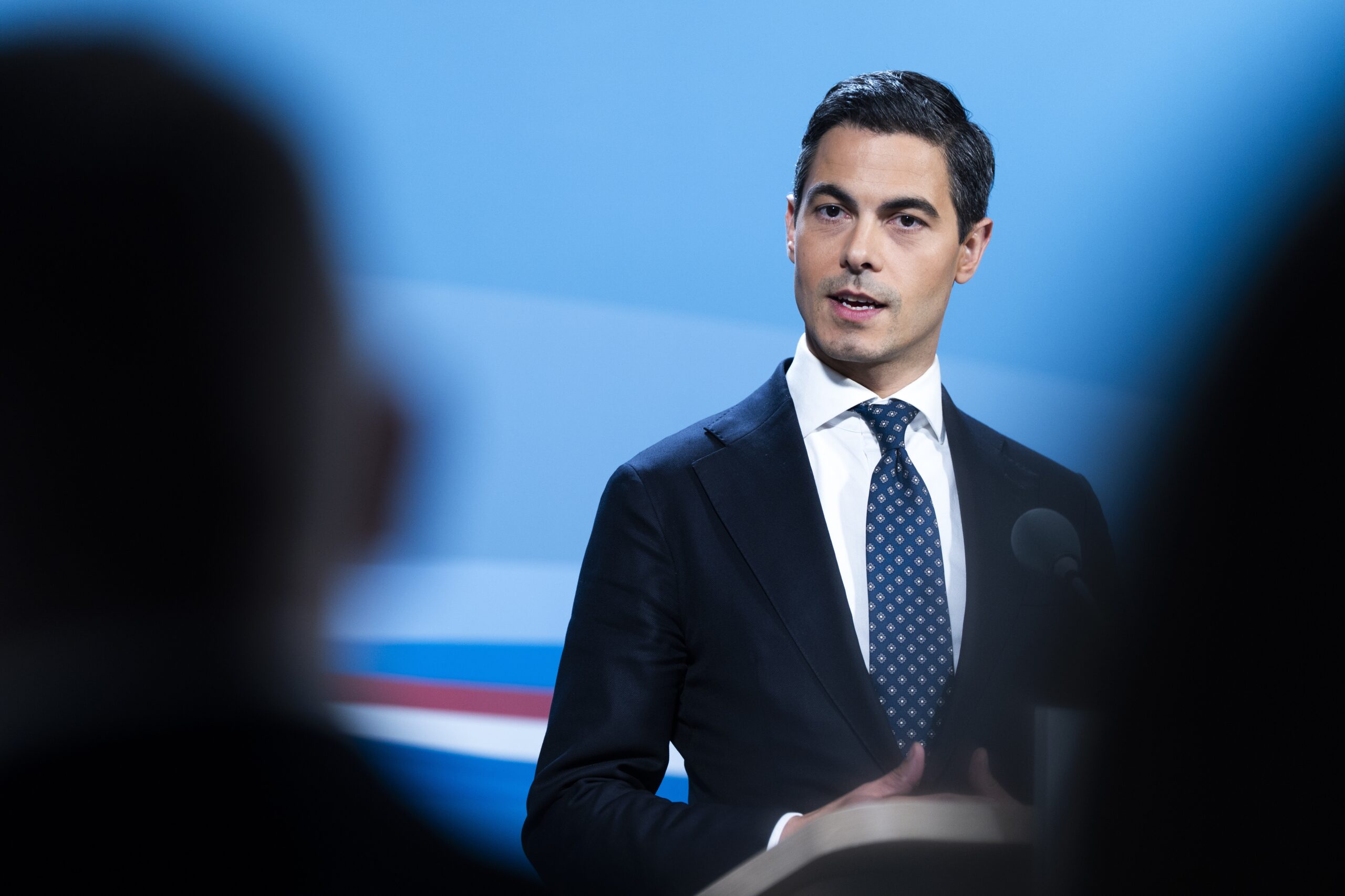 Rob Jetten spreekt bij een podium voor een blauwe achtergrond.