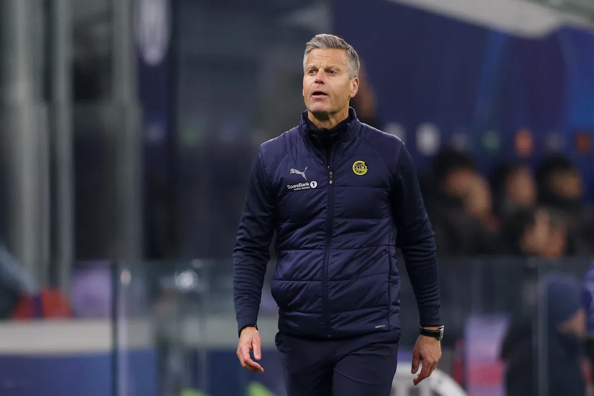 Een voetbaltrainer in donker trainingsjack loopt langs de zijlijn tijdens een wedstrijd.