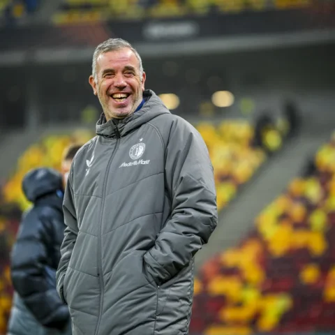 Een lachende voetbaltrainer in een grijze jas langs het veld in een stadion.