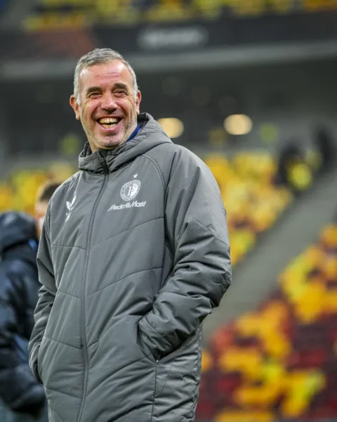 Een lachende voetbaltrainer in een grijze jas langs het veld in een stadion.