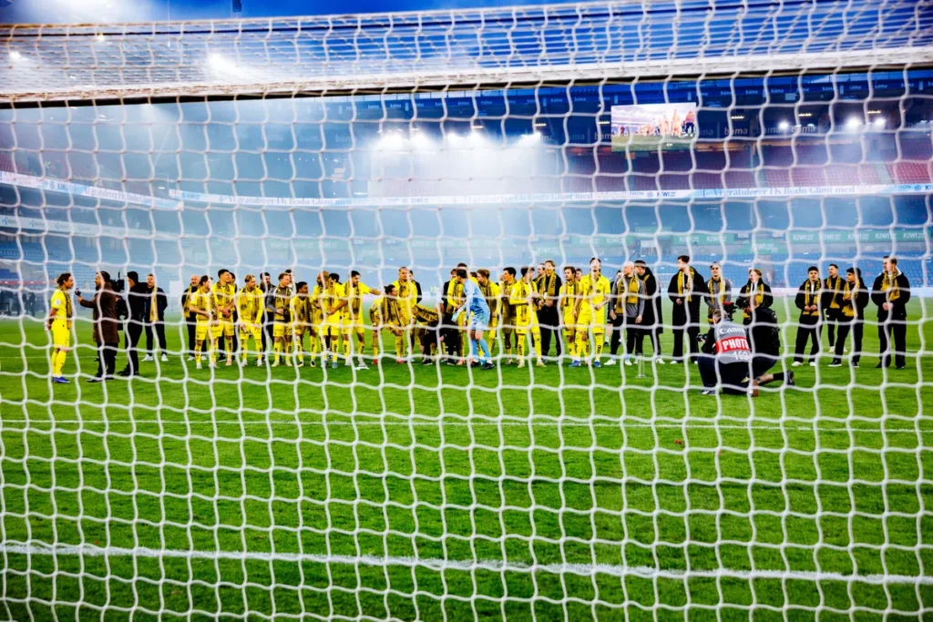 Bodø/Glimt-spelers staan op het veld in het Aspmyra Stadion, gezien door het doelnet.