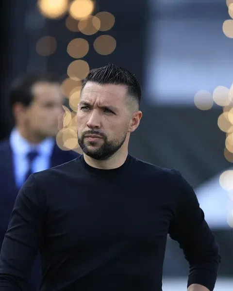 Francesco Farioli staat bij een voetbalwedstrijd met een donkere outfit en een gefocuste blik.