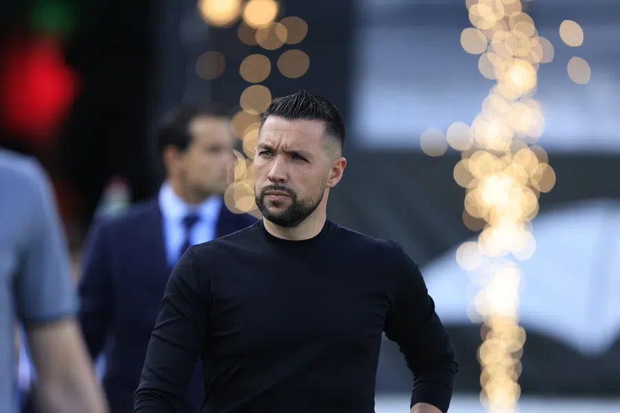 Francesco Farioli staat bij een voetbalwedstrijd met een donkere outfit en een gefocuste blik.