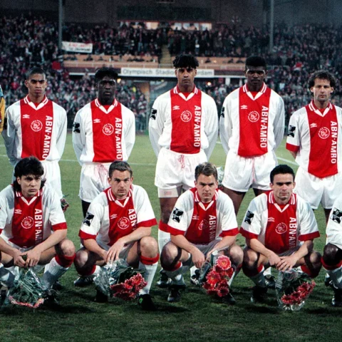 Ajax-selectie poseert voor een groepsfoto in het stadion, met spelers in rood-witte shirts en de doelman links.