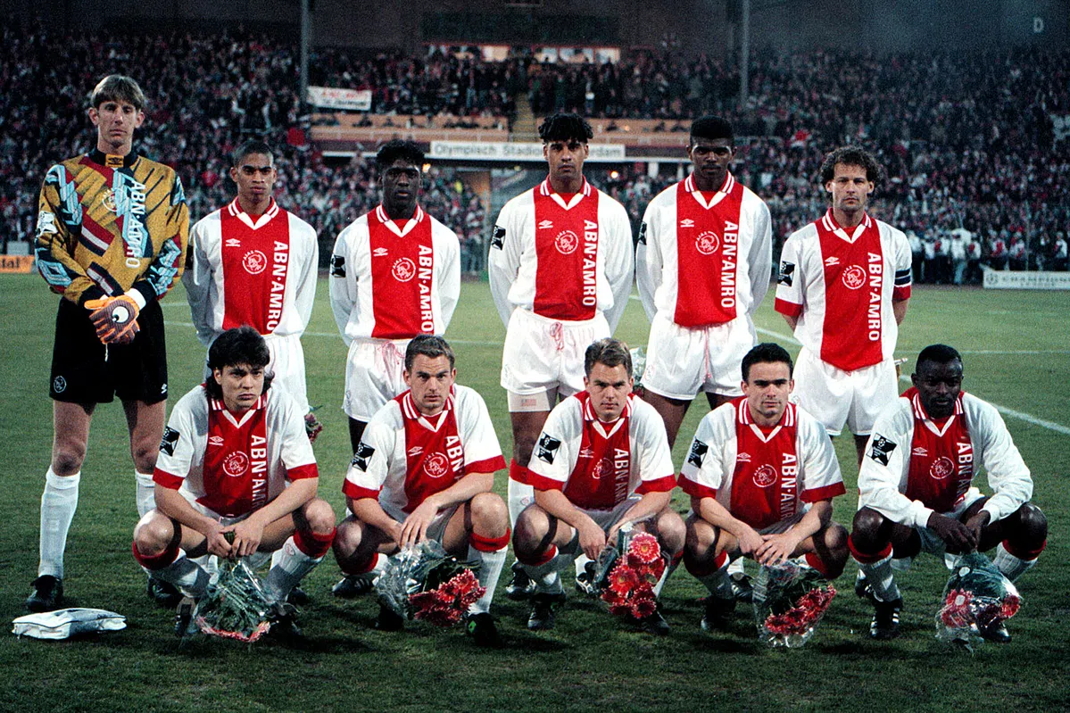 Ajax-selectie poseert voor een groepsfoto in het stadion, met spelers in rood-witte shirts en de doelman links.