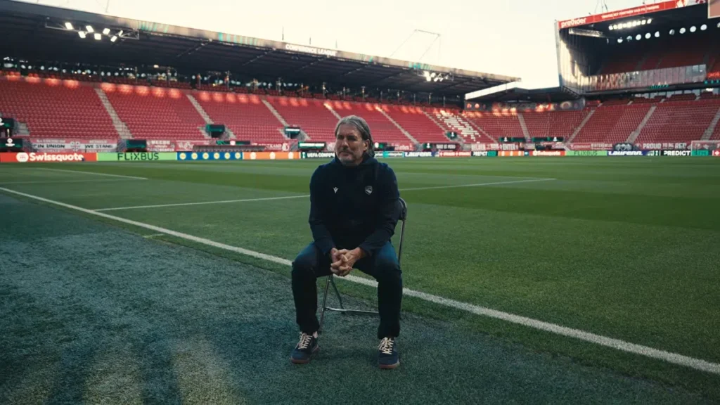 Andres Gerber zit op een inklapstoel op het veld in een leeg voetbalstadion.