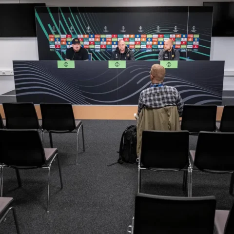 Een vrijwel lege persconferentieruimte met drie mannen aan tafel en één journalist op de voorgrond.