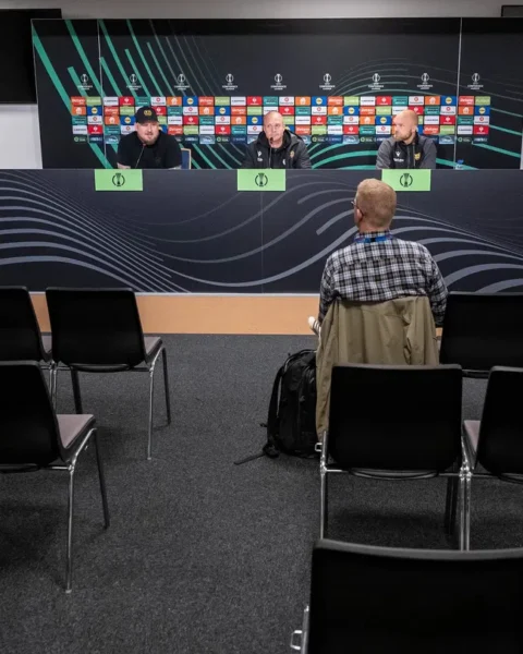 Een vrijwel lege persconferentieruimte met drie mannen aan tafel en één journalist op de voorgrond.