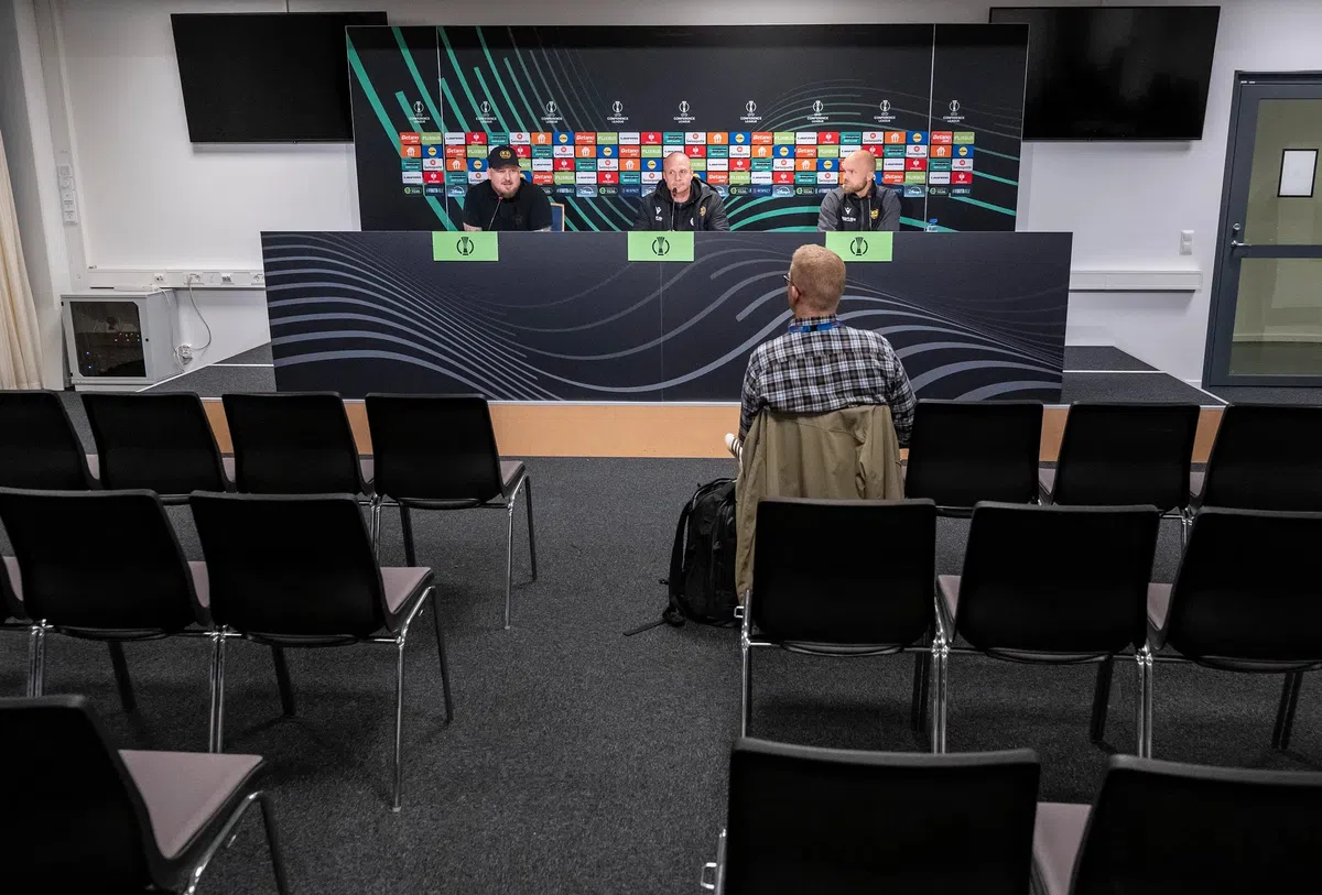 Een vrijwel lege persconferentieruimte met drie mannen aan tafel en één journalist op de voorgrond.
