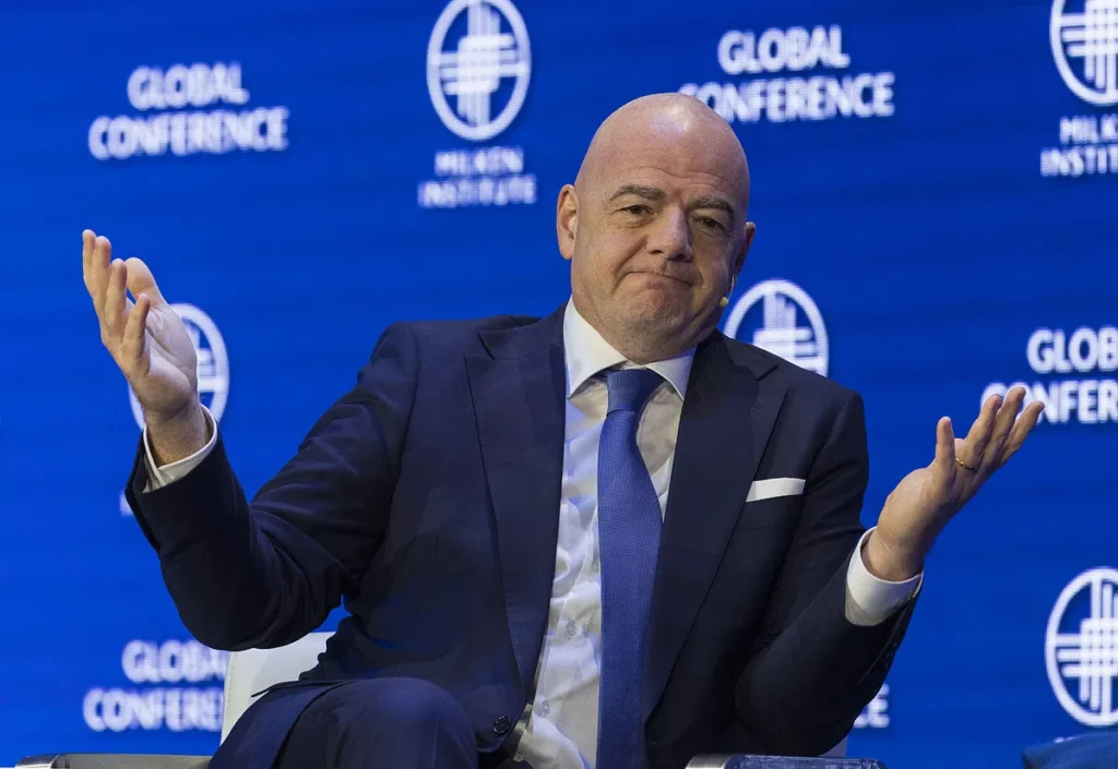 Gianni Infantino zit op een podium met een blauw pak en gespreide handen voor een FIFA Global Conference-achtergrond.