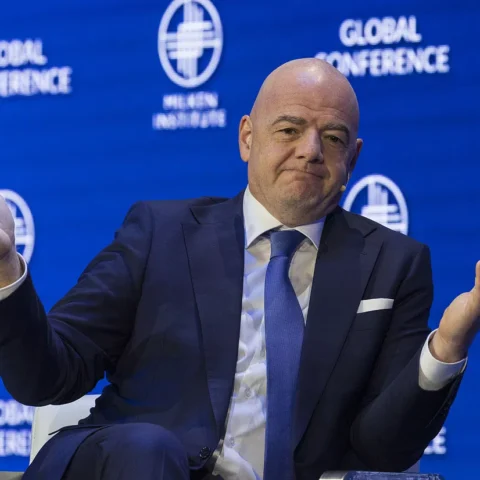 Gianni Infantino zit op een podium met een blauw pak en gespreide handen voor een FIFA Global Conference-achtergrond.