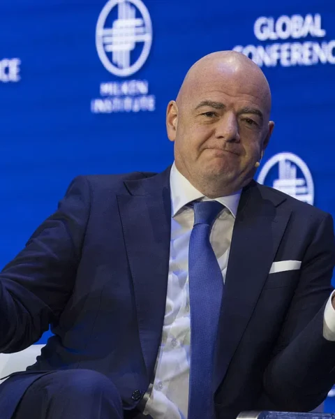 Gianni Infantino zit op een podium met een blauw pak en gespreide handen voor een FIFA Global Conference-achtergrond.